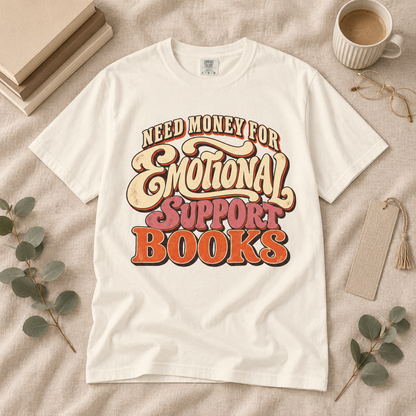 A white graphic T-shirt laid flat on a beige fabric surface with styled props including books, a cup of coffee, glasses, eucalyptus leaves, and a tassel tag. The shirt has a large retro-style text graphic reading “Need Money for Emotional Support Books” in cream, tan, pink, and orange tones.