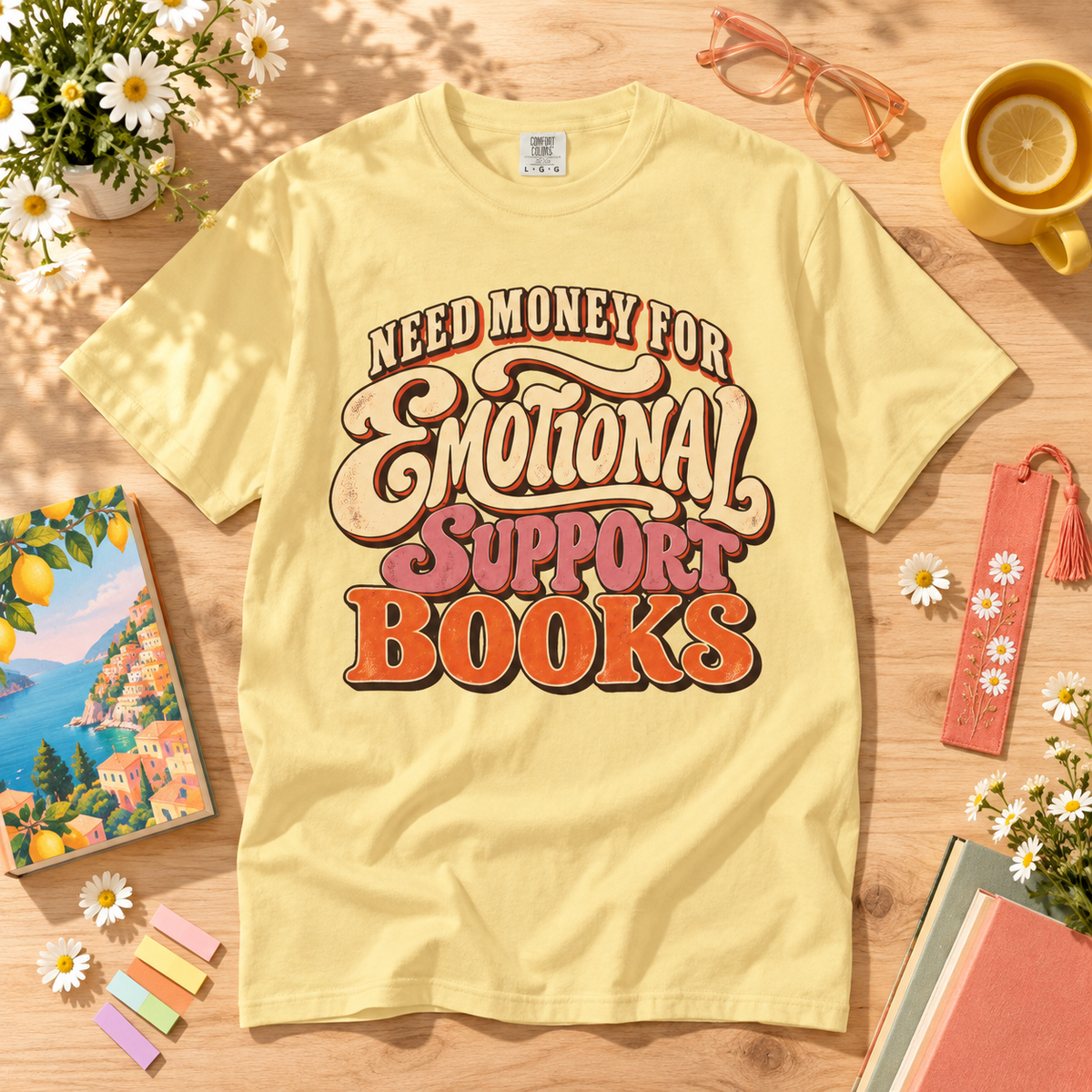 A pale yellow graphic T-shirt laid flat on a wooden surface with styled props including flowers, glasses, a mug, a book, sticky notes, and a bookmark. The shirt reads “NEED MONEY FOR Emotional Support Books” in large retro-style lettering.