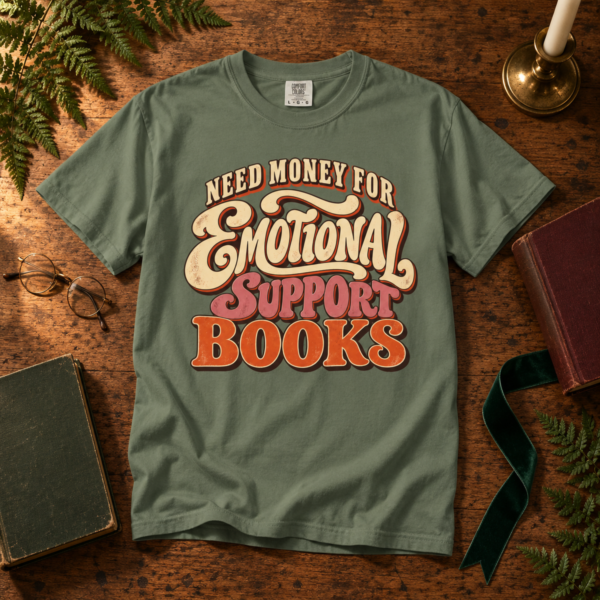 An olive green graphic t-shirt laid flat on a rustic wooden table, surrounded by vintage books, ferns, and a candle. The shirt features a retro-style graphic that reads 'NEED MONEY FOR Emotional Support BOOKS' in cream, pink, and orange lettering.