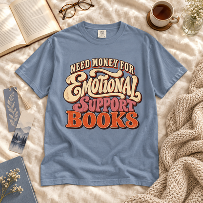 A light blue graphic T-shirt laid flat on a cream fabric surface with cozy props around it, including an open book, glasses, a cup of coffee, a small vase, and a knitted blanket. The shirt has a large retro-style text graphic reading “Need Money for Emotional Support Books.