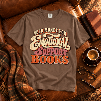 A brown graphic t-shirt laid flat on a brown leather armchair, surrounded by cozy props including a plaid throw blanket, a cup of coffee, eyeglasses, and a pen. The shirt features a retro-style text graphic that reads 'NEED MONEY FOR Emotional SUPPORT BOOKS'.