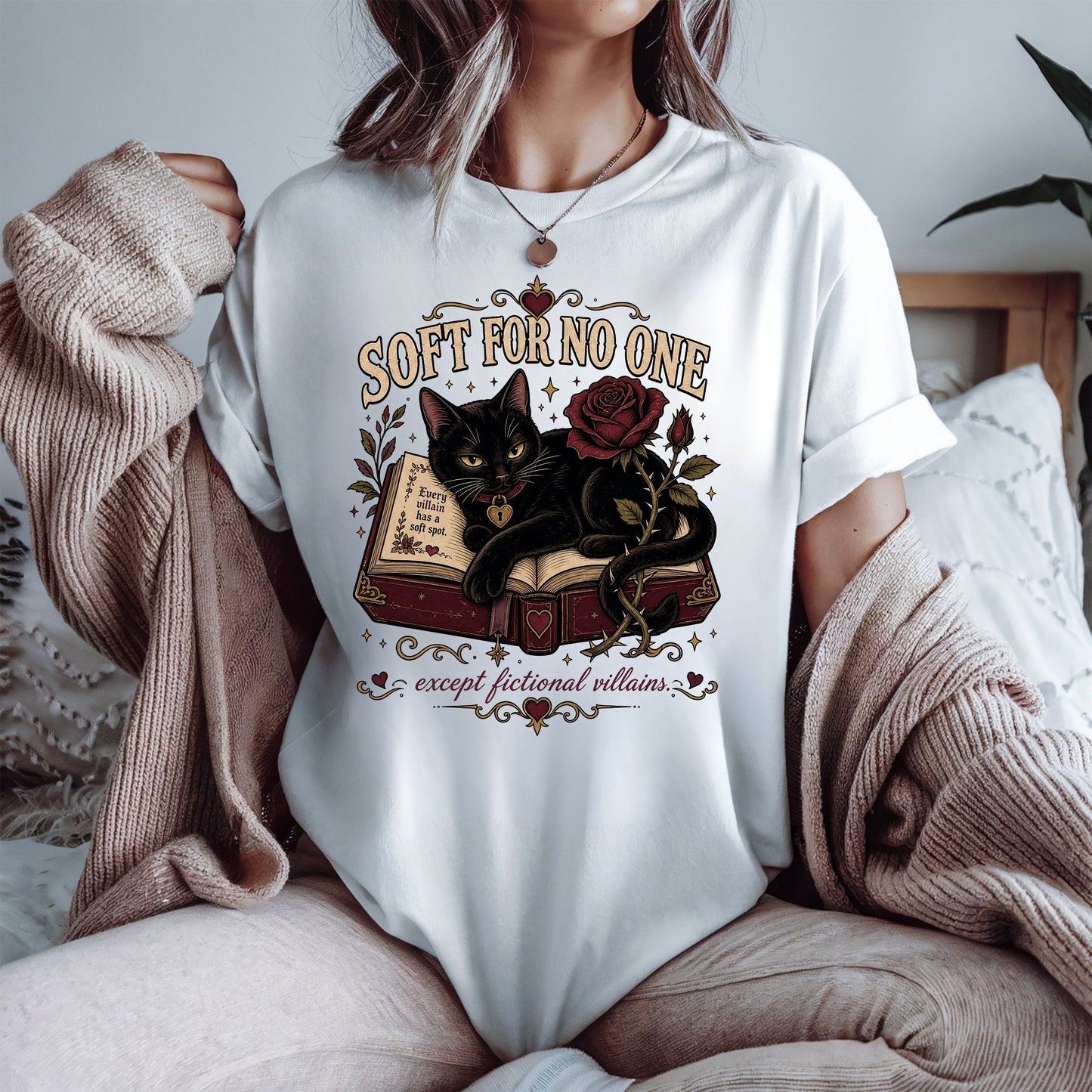 A woman wearing a white graphic t-shirt with a black cat and rose design, styled with a beige knit cardigan in a cozy bedroom setting.