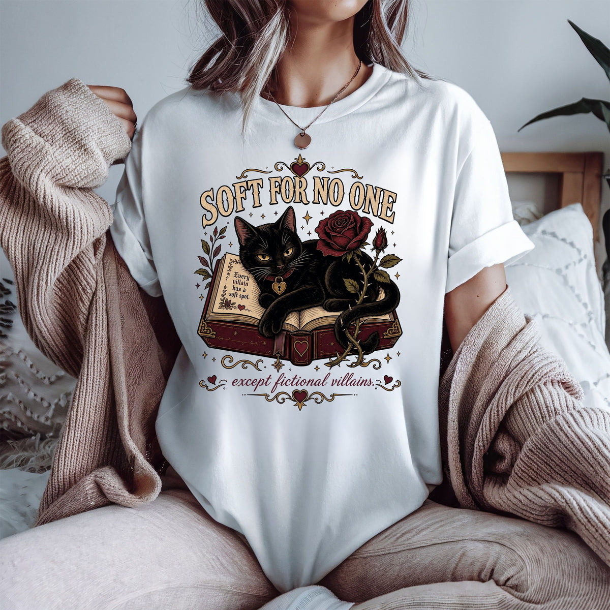 A woman wearing a white graphic t-shirt with a black cat and rose design, styled with a beige knit cardigan in a cozy bedroom setting.