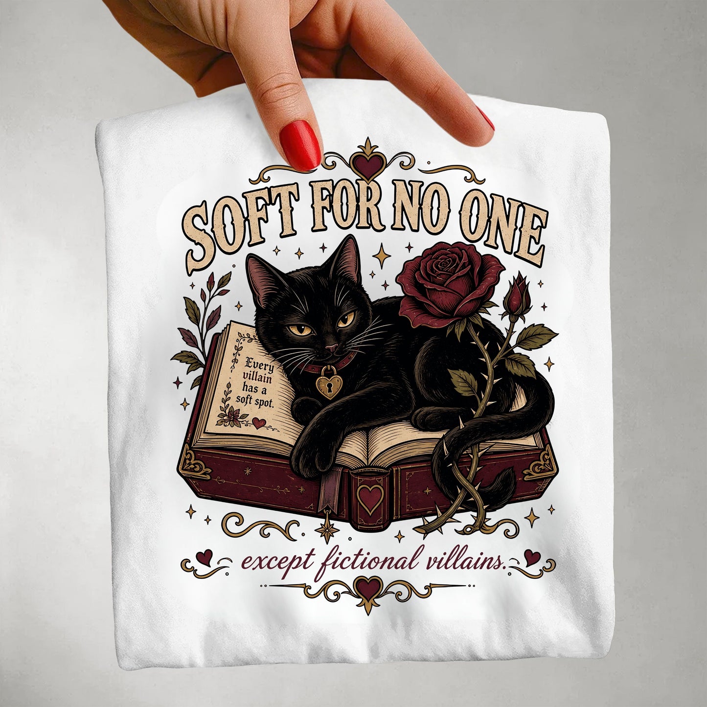 A hand with red nail polish holds up a white graphic t-shirt. The shirt features a gothic-style illustration of a black cat lying on an open book next to a dark red rose, with the text 'SOFT FOR NO ONE' and 'except fictional villains'.