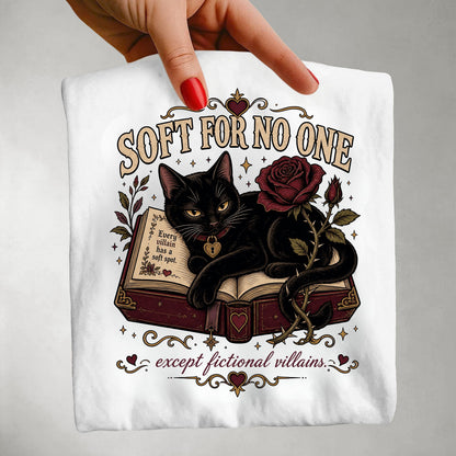 A hand with red nail polish holds up a white graphic t-shirt. The shirt features a gothic-style illustration of a black cat lying on an open book next to a dark red rose, with the text 'SOFT FOR NO ONE' and 'except fictional villains'.