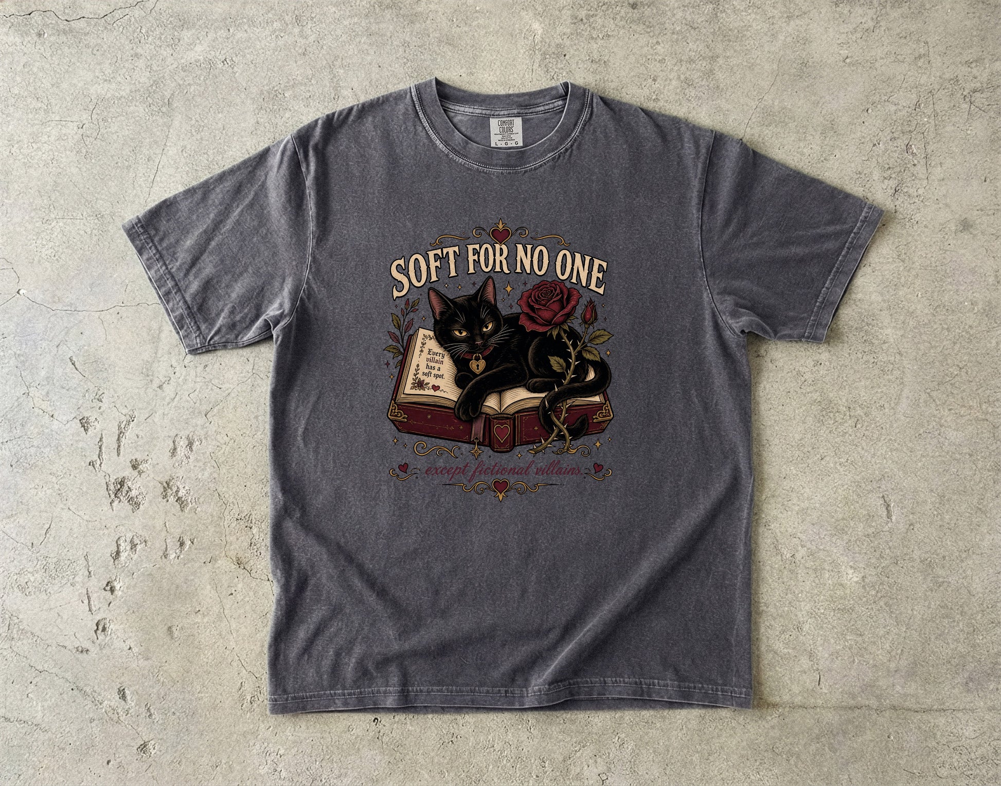 A dark gray graphic t-shirt laid flat on a textured concrete surface. The shirt features a graphic of a black cat resting on an open book next to a red rose, with the text 'SOFT FOR NO ONE' arched above the illustration.
