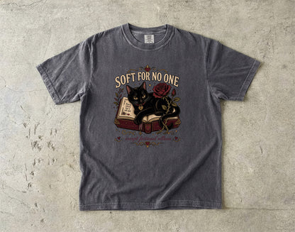 A dark gray graphic t-shirt laid flat on a textured concrete surface. The shirt features a graphic of a black cat resting on an open book next to a red rose, with the text 'SOFT FOR NO ONE' arched above the illustration.