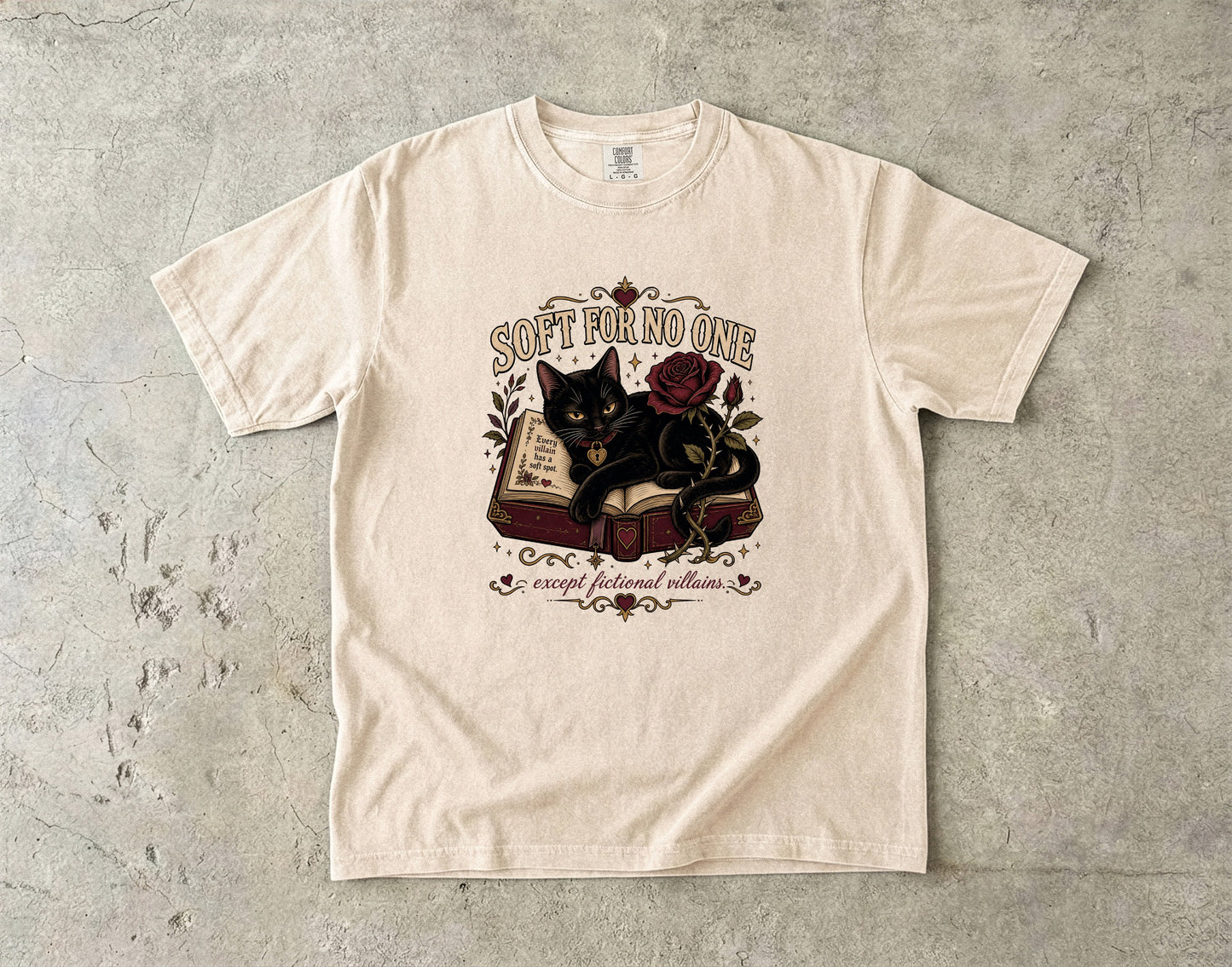 A cream-colored short-sleeve t-shirt with a vintage-style graphic of a black cat on an open book with a red rose, laid flat on a textured gray concrete surface. The graphic includes the text 'SOFT FOR NO ONE' and 'except for my faithful cat'.