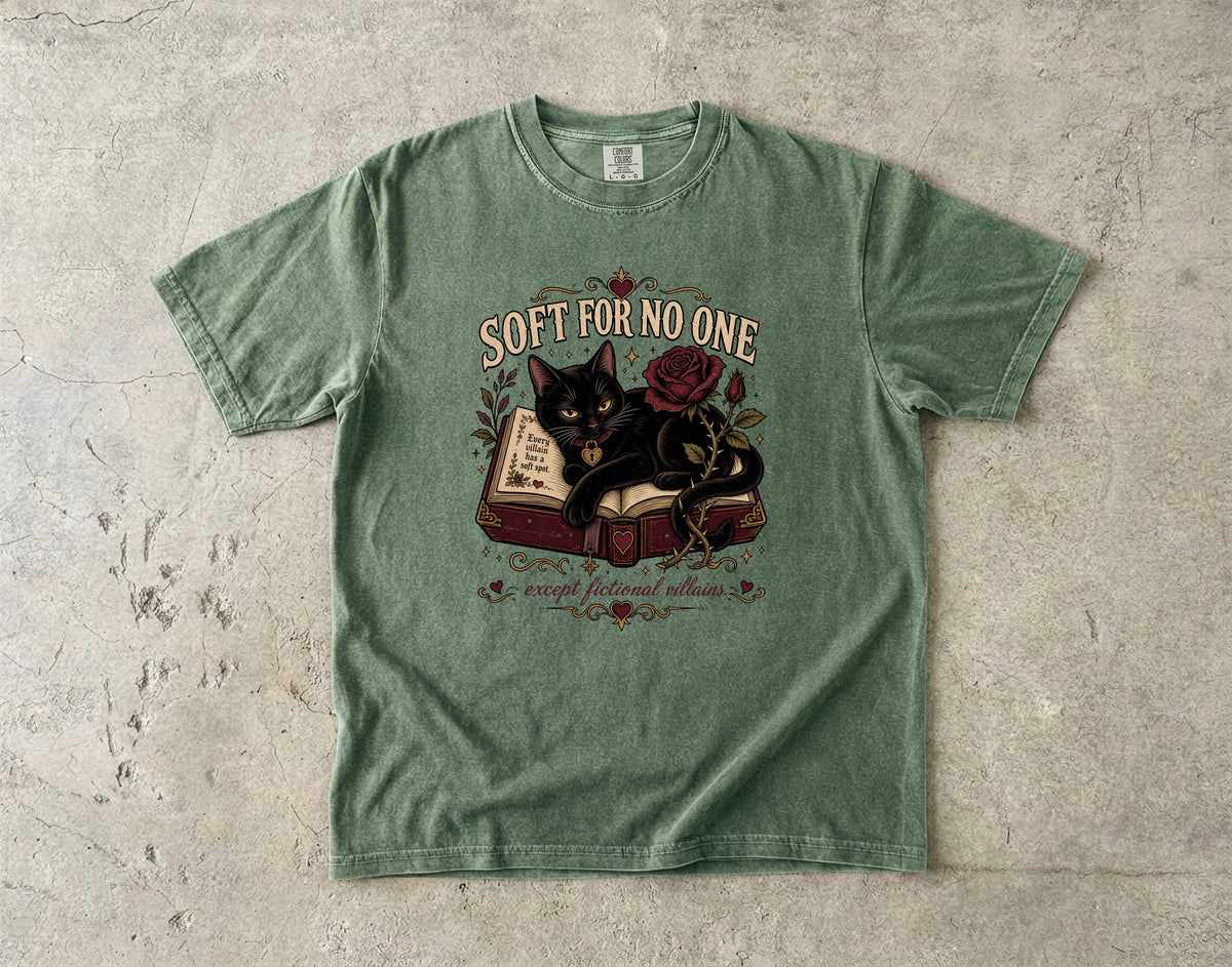 A sage green graphic t-shirt laid flat on a textured concrete surface. The shirt features a central graphic of a black cat sitting on an open book next to a red rose, with the text 'SOFT FOR NO ONE' above and 'escape fictional reality' below.