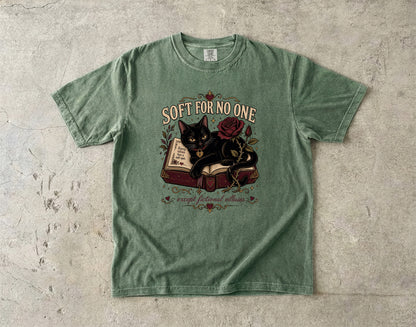 A sage green graphic t-shirt laid flat on a textured concrete surface. The shirt features a central graphic of a black cat sitting on an open book next to a red rose, with the text 'SOFT FOR NO ONE' above and 'escape fictional reality' below.