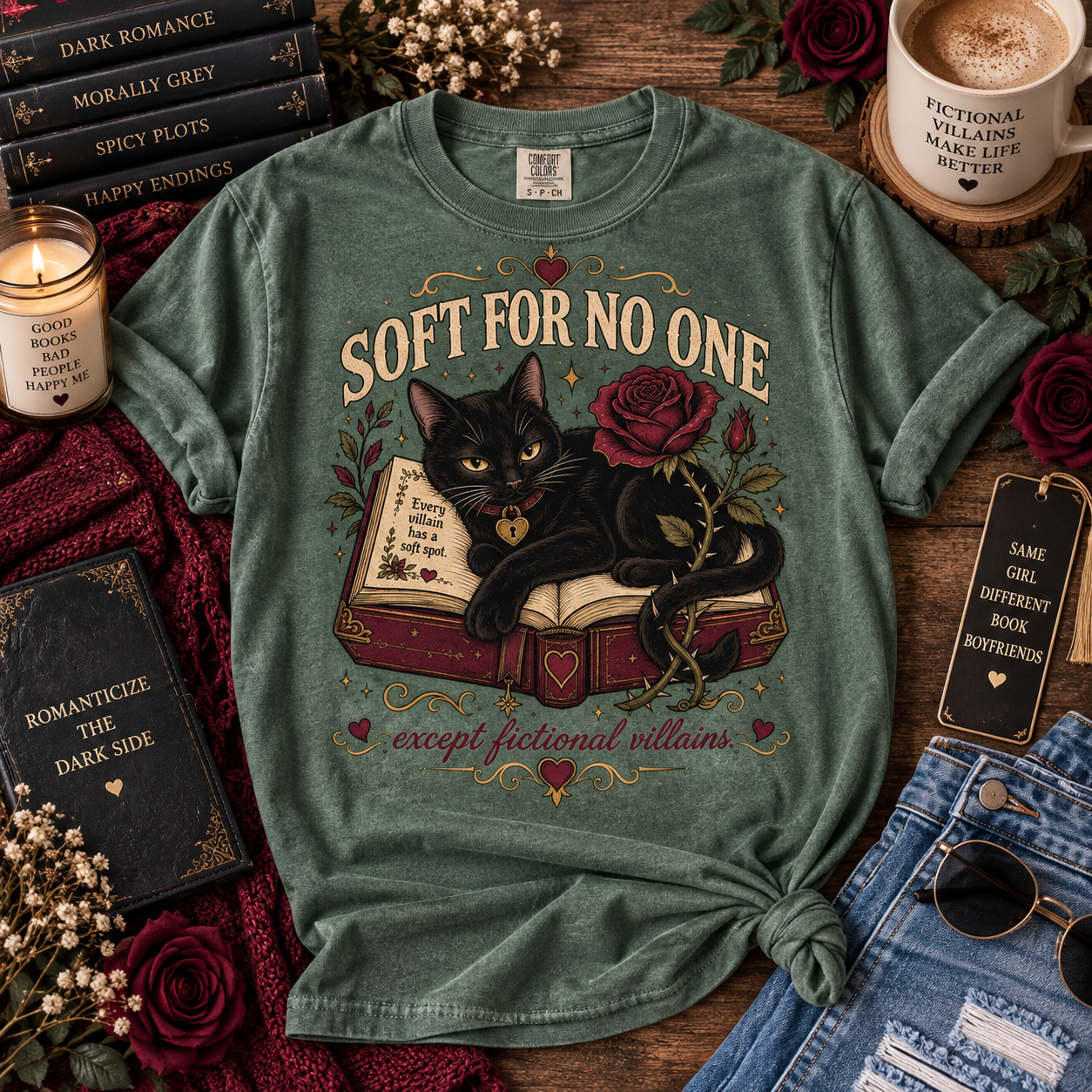 A green graphic t-shirt featuring a black cat on an open book with roses, surrounded by cozy styled props like books, a candle, sunglasses, and denim jeans on a wooden surface.
