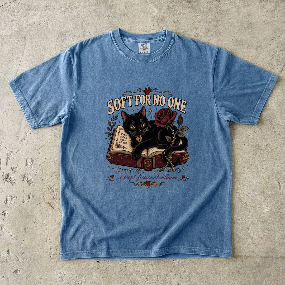 A light blue short-sleeve graphic t-shirt laid flat on a textured concrete surface. The graphic features a black cat curled up on an open book next to a red rose, with the text 'SOFT FOR NO ONE' above and 'except for you villains' below.