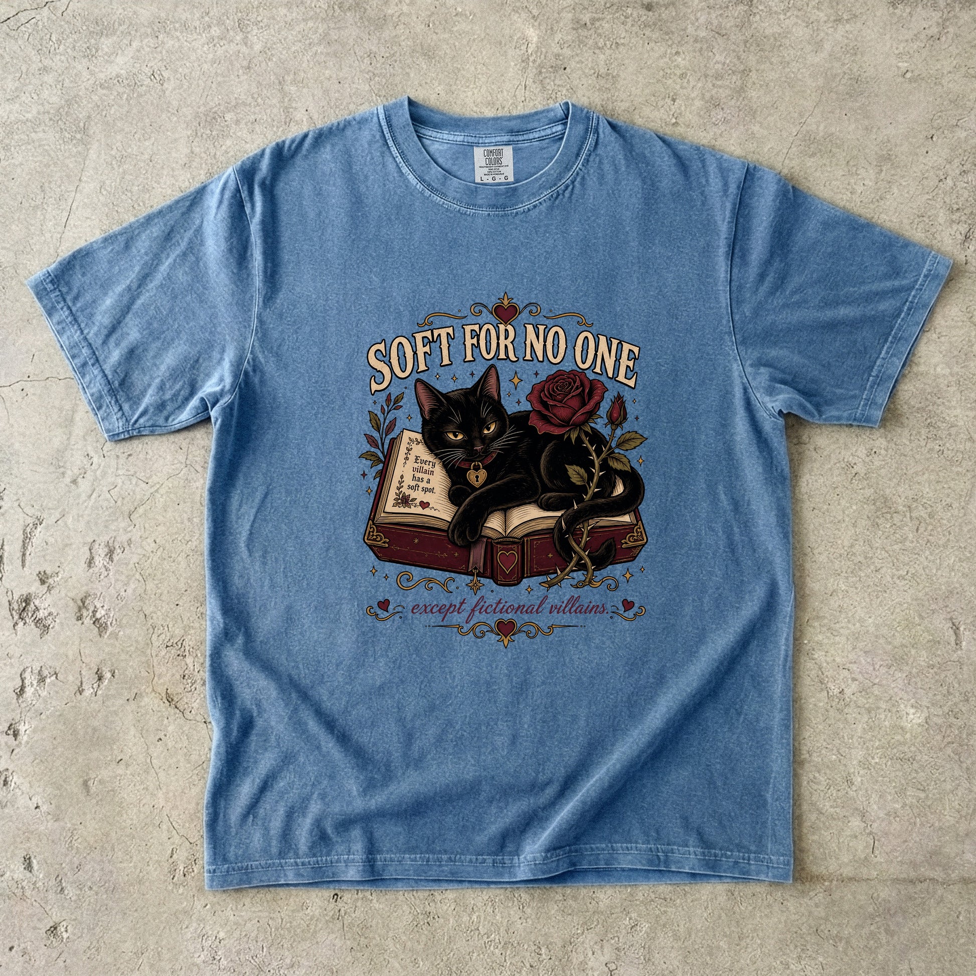 A light blue short-sleeve graphic t-shirt laid flat on a textured concrete surface. The graphic features a black cat curled up on an open book next to a red rose, with the text 'SOFT FOR NO ONE' above and 'except for you villains' below.