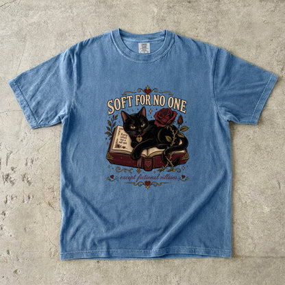 A light blue short-sleeve graphic t-shirt laid flat on a textured concrete surface. The graphic features a black cat curled up on an open book next to a red rose, with the text 'SOFT FOR NO ONE' above and 'except for you villains' below.