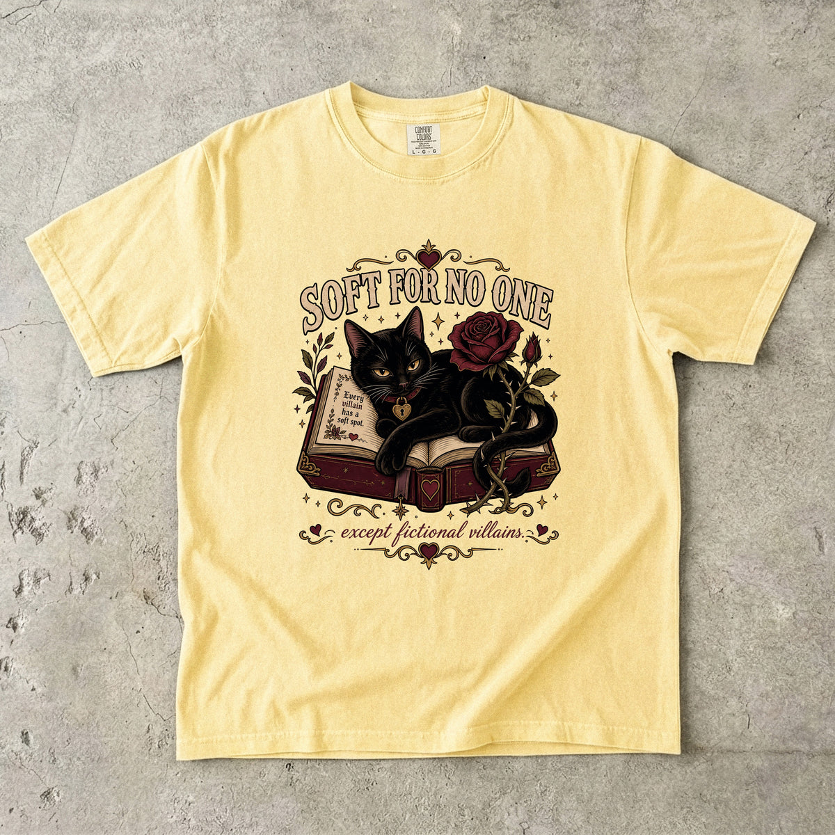 A pale yellow graphic t-shirt laid flat on a textured gray concrete background. The shirt features a central graphic of a black cat curled up on an open book next to a red rose, with the text 'SOFT FOR NO ONE' above and 'except fictional villains' below.