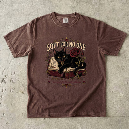 A brown short-sleeve graphic t-shirt laid flat on a textured concrete surface. The graphic features a black cat lying on an open book next to a red rose, with the text 'SOFT FOR NO ONE' above and 'heart faithful readers' below.