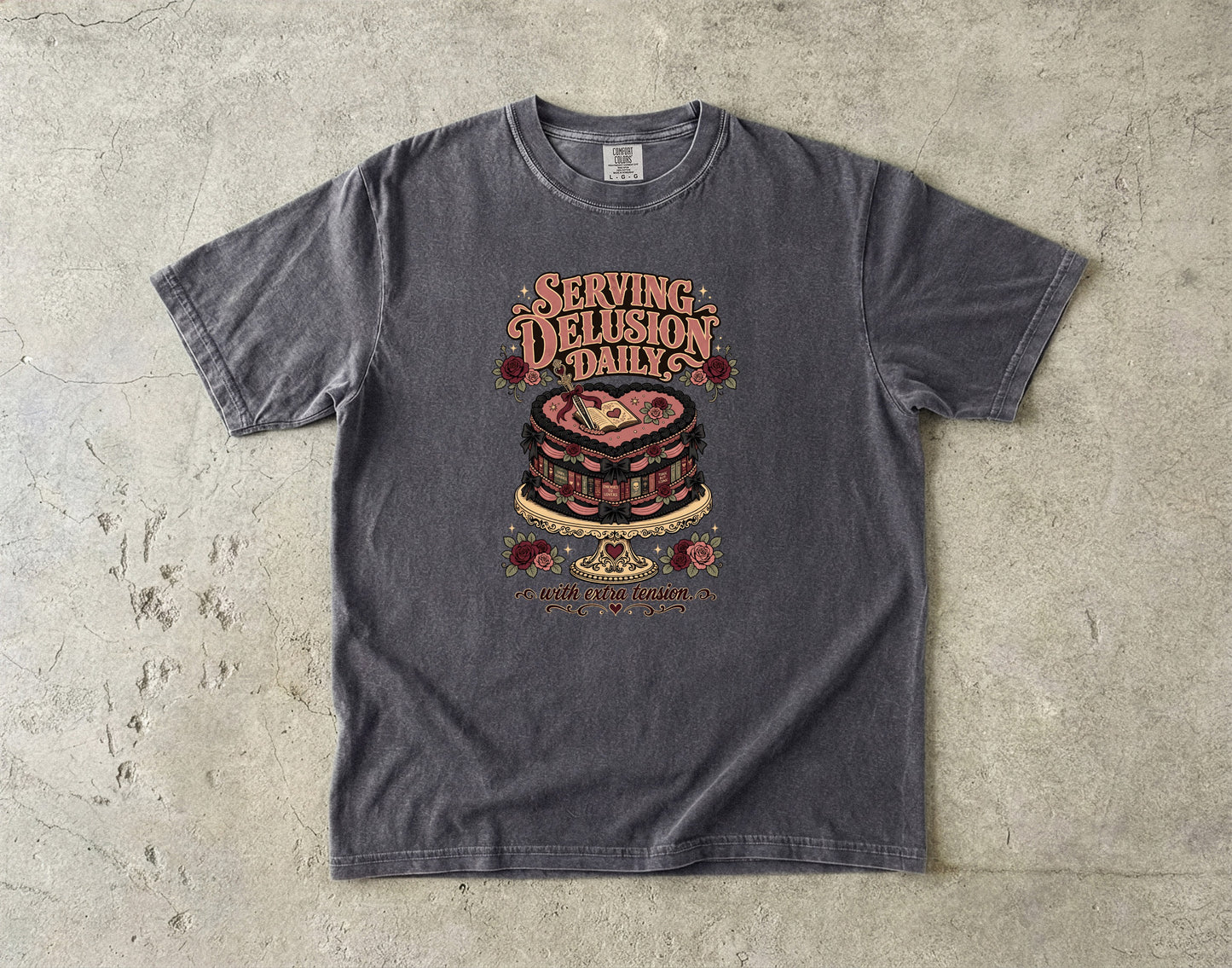 A dark gray graphic t-shirt laid flat on a textured concrete surface. The shirt features a vintage-style graphic of a layered cake with the text 'SERVING DELUSION DAILY' and 'NOT A NORMAL HUMAN' printed around it.