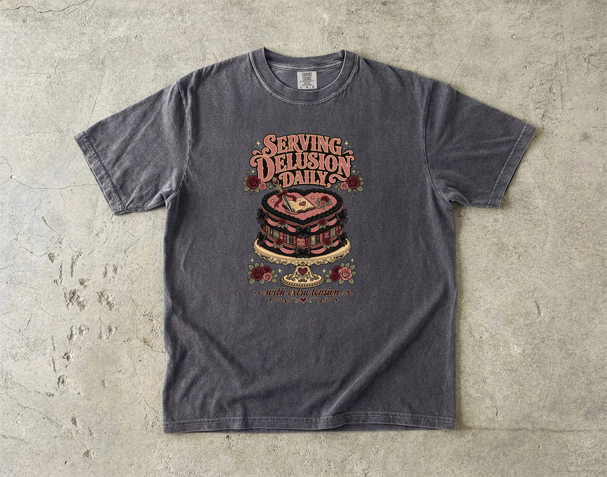 A dark gray graphic t-shirt laid flat on a textured concrete surface. The shirt features a vintage-style graphic of a layered cake with the text 'SERVING DELUSION DAILY' and 'NOT A NORMAL HUMAN' printed around it.