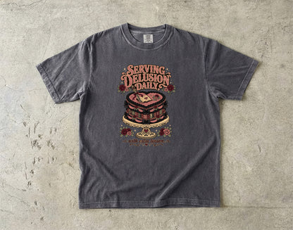 A dark gray graphic t-shirt laid flat on a textured concrete surface. The shirt features a vintage-style graphic of a layered cake with the text 'SERVING DELUSION DAILY' and 'NOT A NORMAL HUMAN' printed around it.