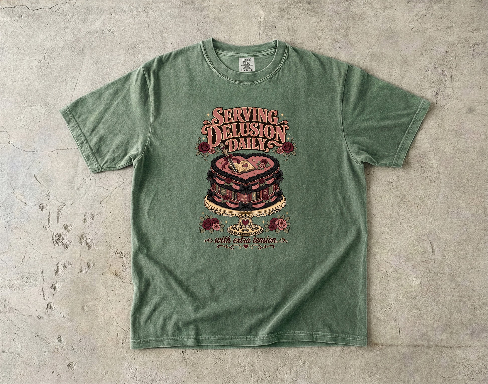 A sage green short-sleeve t-shirt with a vintage-style graphic of a layered chocolate cake and the text 'SERVING DELUSION DAILY' laid flat on a textured concrete surface.