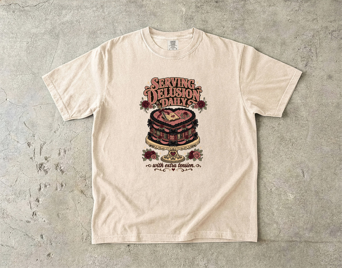 A cream-colored short-sleeve t-shirt with a vintage-style graphic of a layered cake and the text 'Serving Delusion Daily' and 'with extra icing', laid flat on a textured gray concrete surface.