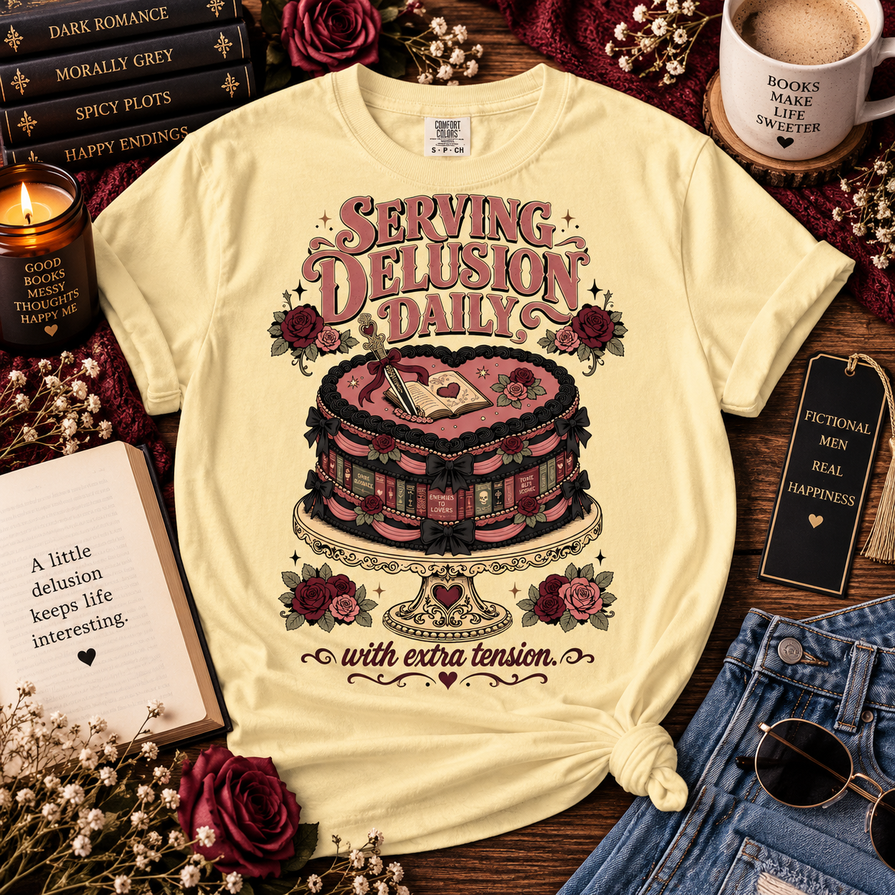 A pale yellow graphic T-shirt laid flat on a rustic wood surface, surrounded by decorative props including books, a candle, an open book, dried flowers, denim jeans, and a mug. The shirt has a large vintage-style illustration of a pink and black layered cake with roses and the text 'Serving Delusion Daily with extra tension.