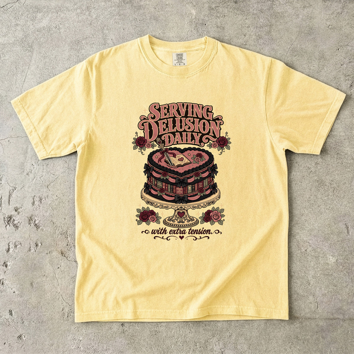A pale yellow graphic t-shirt laid flat on a textured gray concrete surface. The graphic features a vintage-style illustration of a pink and red heart-shaped cake with the text 'SERVING DELUSION DAILY' above it and 'with eternal vision.