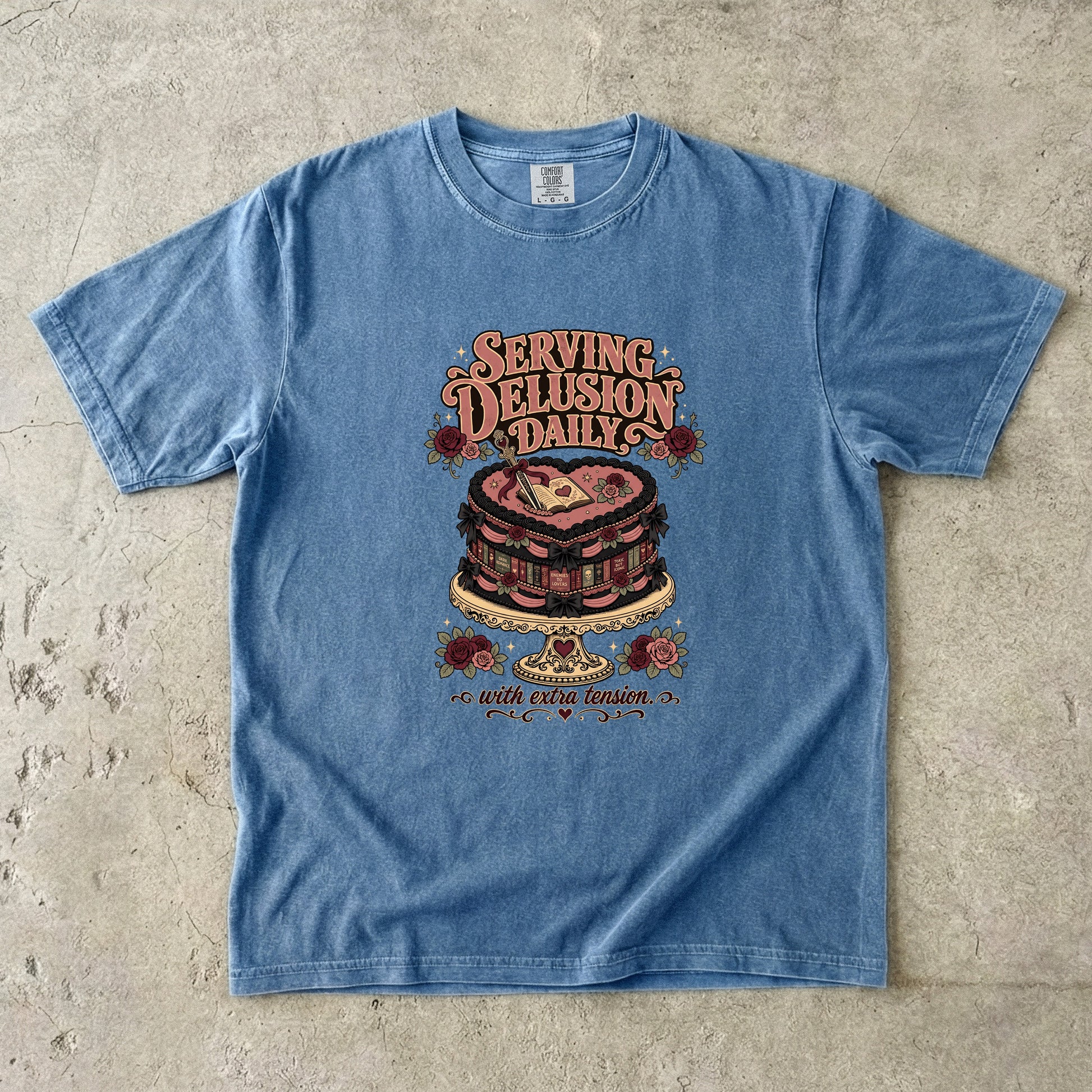 A light blue washed graphic t-shirt laid flat on a textured concrete surface. The shirt features a vintage-style graphic of a heart-shaped cake on a stand with the text 'SERVING DELUSION DAILY' and 'with evil vision' below it.