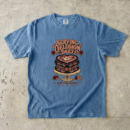 A light blue washed graphic t-shirt laid flat on a textured concrete surface. The shirt features a vintage-style graphic of a heart-shaped cake on a stand with the text 'SERVING DELUSION DAILY' and 'with evil vision' below it.