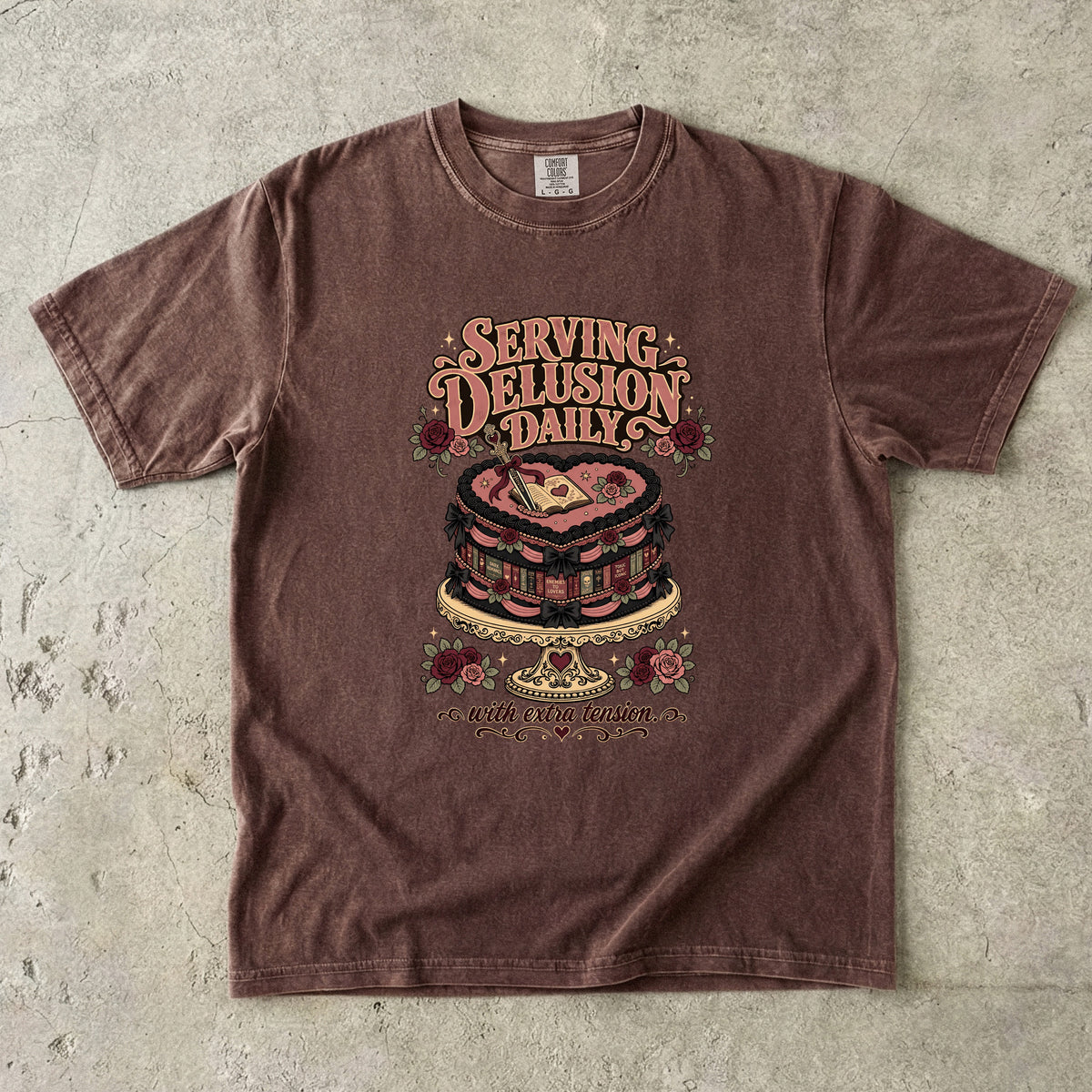 A brown short-sleeve graphic t-shirt laid flat on a textured concrete surface. The shirt features a retro-style graphic of a layered cake with a heart on top, surrounded by roses and the text 'SERVING DELUSION DAILY' and 'BUT THE REAL QUESTION IS'.