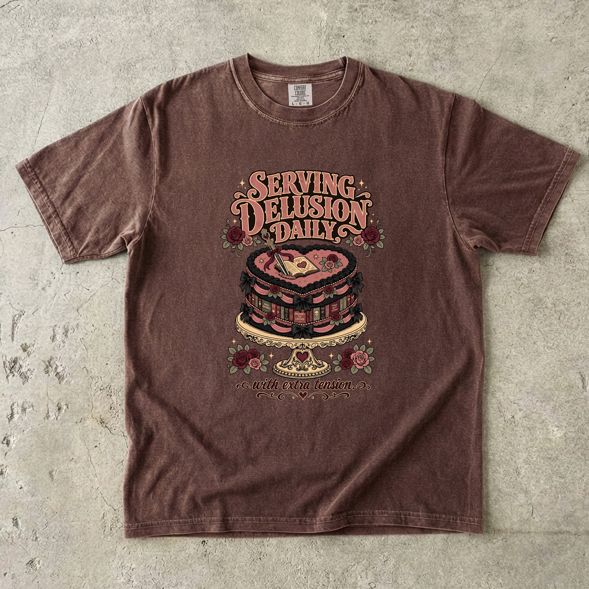 A brown short-sleeve graphic t-shirt laid flat on a textured concrete surface. The shirt features a retro-style graphic of a layered cake with a heart on top, surrounded by roses and the text 'SERVING DELUSION DAILY' and 'BUT THE REAL QUESTION IS'.