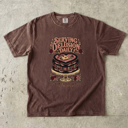A brown short-sleeve graphic t-shirt laid flat on a textured concrete surface. The shirt features a retro-style graphic of a layered cake with a heart on top, surrounded by roses and the text 'SERVING DELUSION DAILY' and 'BUT THE REAL QUESTION IS'.