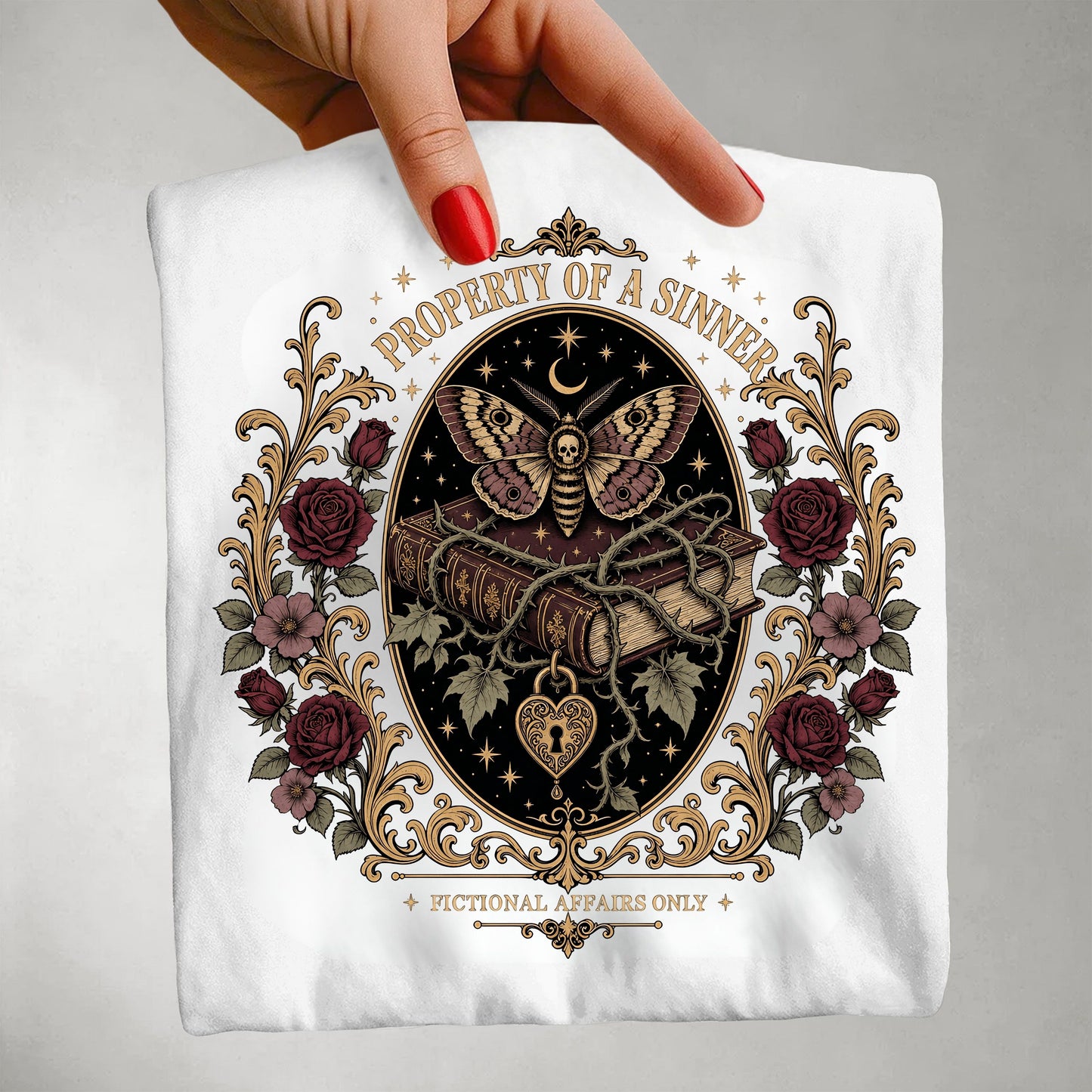 A hand with red nail polish holds a white t-shirt featuring a detailed gothic graphic. The graphic displays a winged demon holding a key, bound by vines, with a crescent moon and stars, surrounded by a frame of dark red roses and gold filigree.