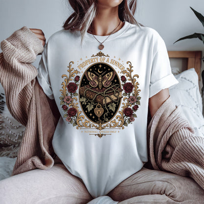 A person wearing a white oversized T-shirt with a large vintage-style butterfly and floral crest graphic on the front, styled with a beige knit sweater draped off the shoulders in a bedroom setting.