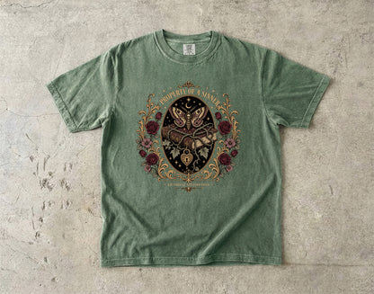 A sage green short-sleeve t-shirt with a detailed gothic graphic of a skeleton hand holding a dagger, surrounded by a butterfly, roses, and the text 'PROPERLY UNARMED' on a textured concrete background.