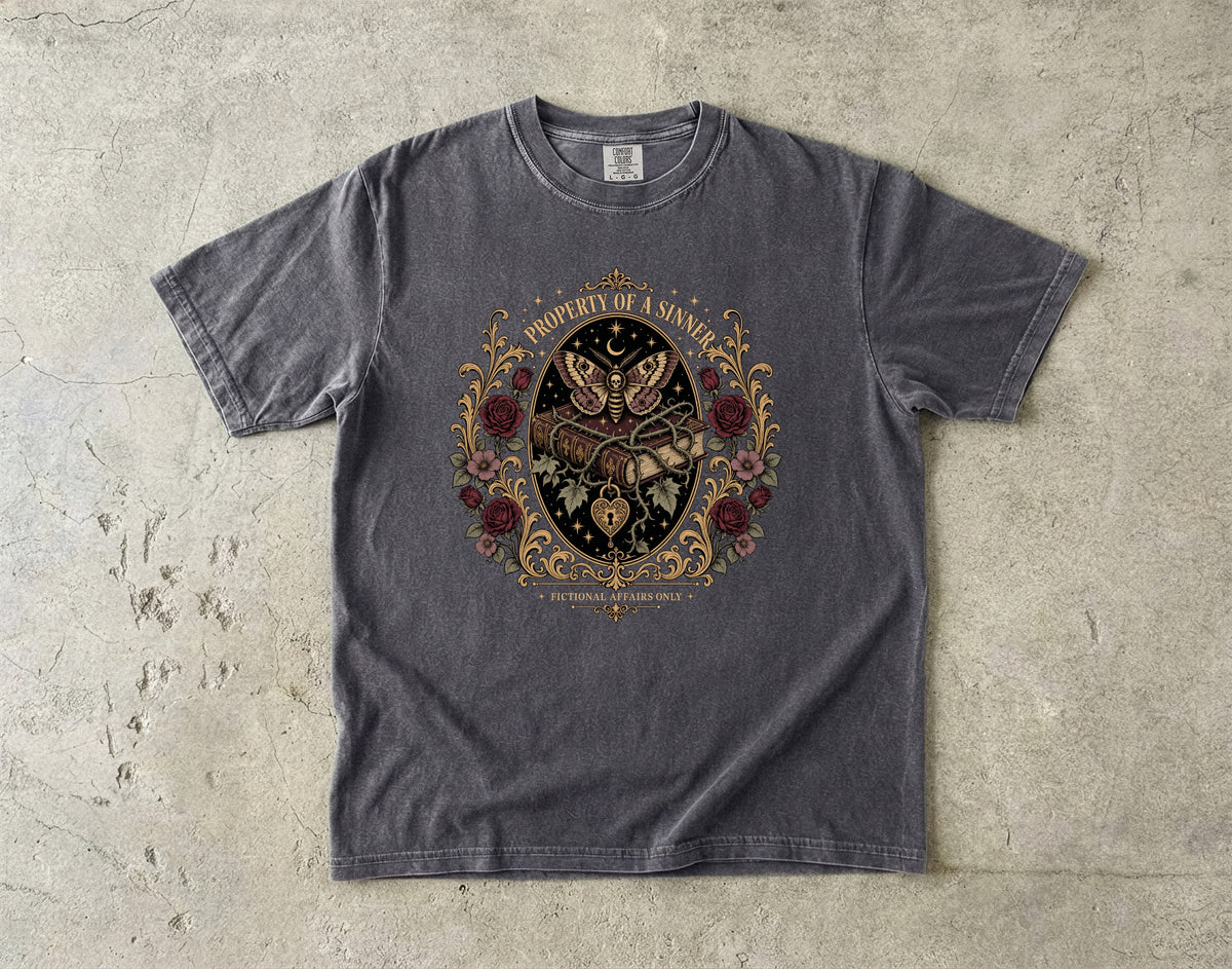 A dark gray heather crewneck t-shirt with a detailed graphic print on the front, laid flat on a textured concrete surface. The graphic features a central black shield with a butterfly, surrounded by red roses and gold filigree, with the text 'PROPERTY OF A MAN' arched above and 'LUX ORBIS MUNDI' below.