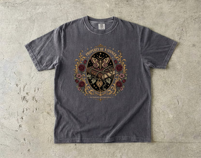 A dark gray heather crewneck t-shirt with a detailed graphic print on the front, laid flat on a textured concrete surface. The graphic features a central black shield with a butterfly, surrounded by red roses and gold filigree, with the text 'PROPERTY OF A MAN' arched above and 'LUX ORBIS MUNDI' below.