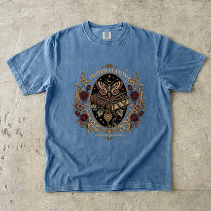 A light blue short-sleeve t-shirt with a detailed gothic graphic print featuring a skeleton, roses, and a crescent moon, laid flat on a textured concrete surface.