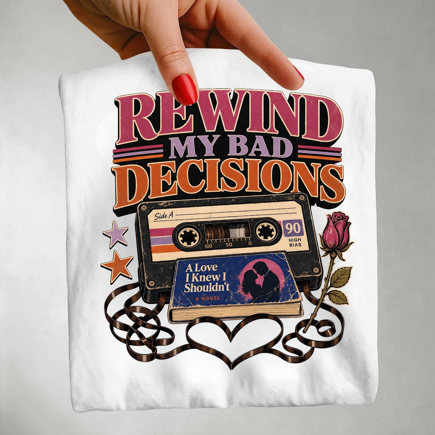 A hand with red nail polish holds up a white t-shirt featuring a retro graphic that reads 'REWIND MY BAD DECISIONS' above a vintage cassette tape, a book, stars, and a rose.