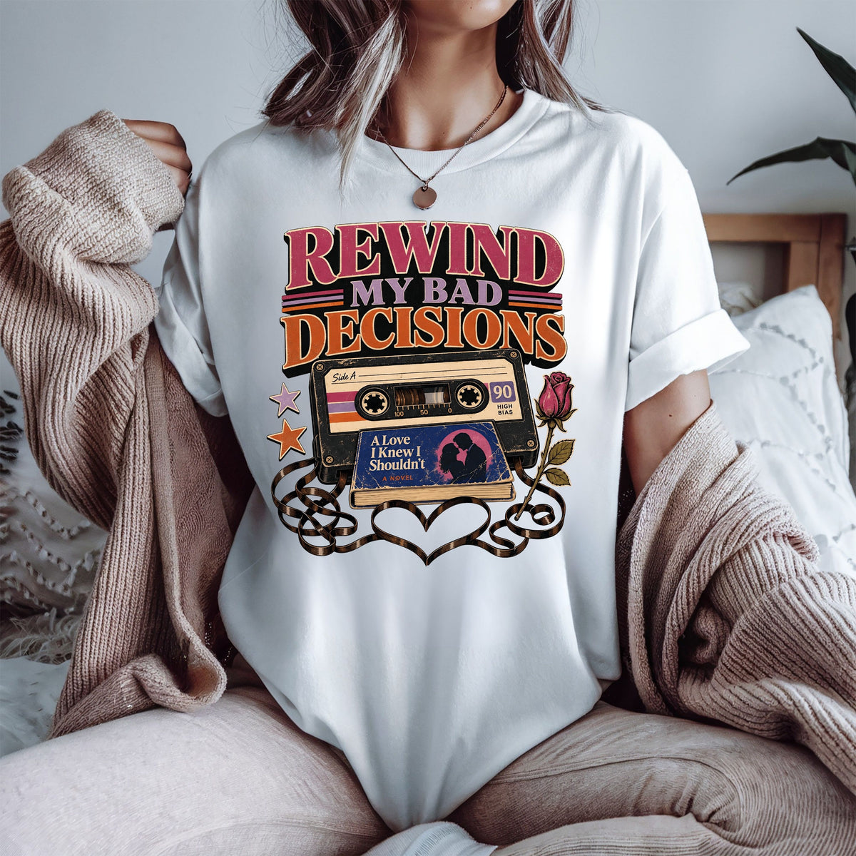 A woman wearing a white graphic t-shirt with a retro cassette tape design and the text 'REWIND MY BAD DECISIONS'. She is sitting in a cozy room with a beige knit sweater draped over her shoulders.