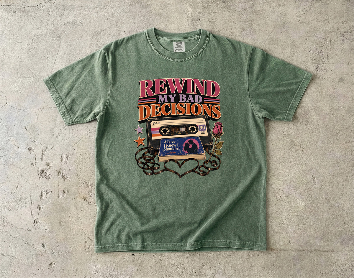 A green graphic t-shirt laid flat on a textured concrete surface. The shirt features a retro cassette tape graphic with the text 'REWIND MY BAD DECISIONS' in pink and orange, and 'I Love Myself' on the tape label.