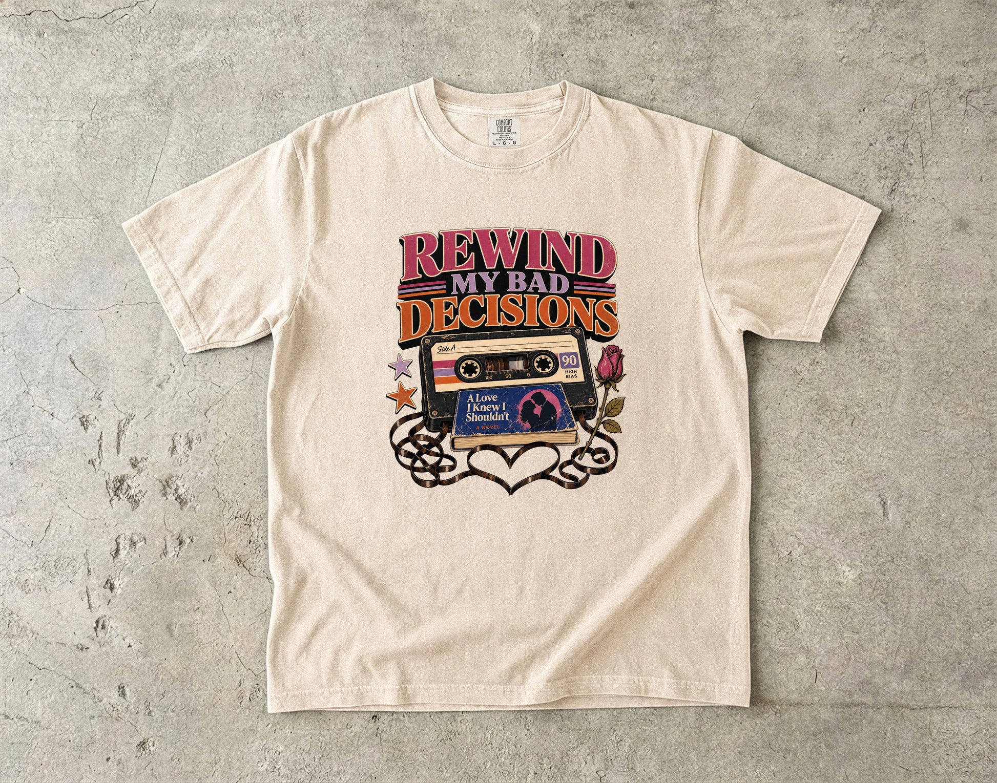 A cream-colored short-sleeve t-shirt with a retro graphic that reads 'REWIND MY BAD DECISIONS' above a cassette tape and heart design, laid flat on a textured gray concrete surface.