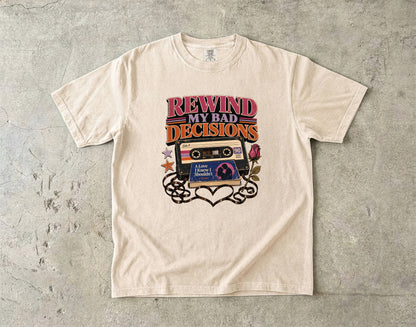 A cream-colored short-sleeve t-shirt with a retro graphic that reads 'REWIND MY BAD DECISIONS' above a cassette tape and heart design, laid flat on a textured gray concrete surface.