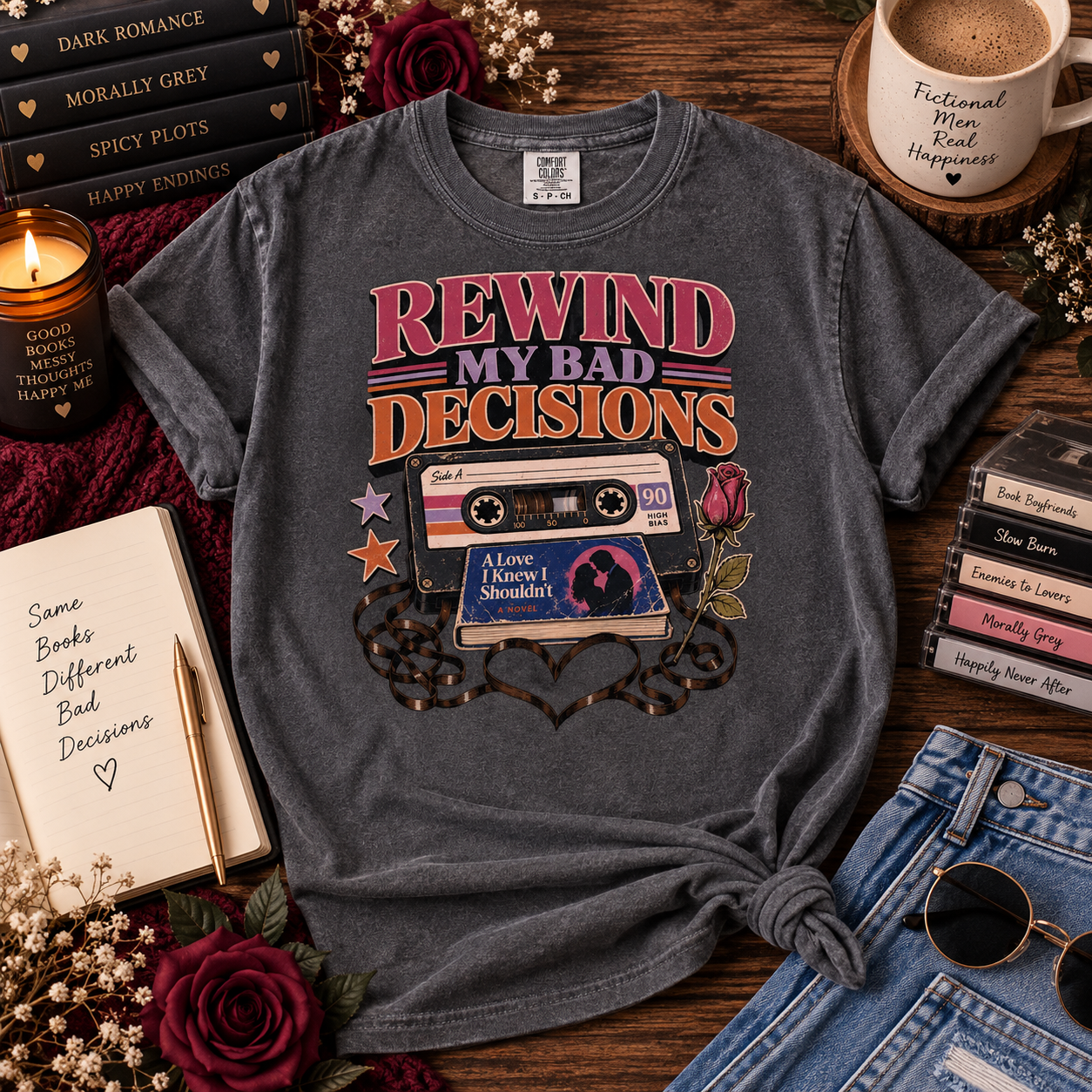 A dark gray graphic t-shirt featuring retro cassette tape and book designs with the text 'REWIND MY BAD DECISIONS'. The shirt is styled in a cozy flat-lay arrangement on a wooden table with a candle, notebook, books, coffee mug, and denim jeans.
