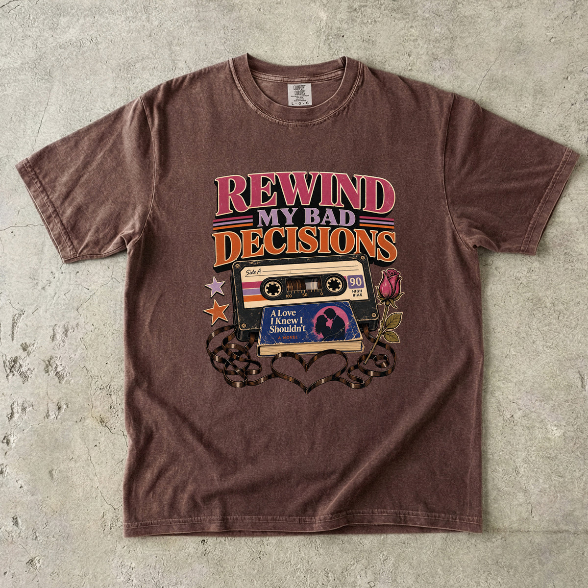A brown short-sleeve crewneck t-shirt laid flat on a textured gray concrete surface. The shirt features a retro graphic of a cassette tape with the text 'REWIND MY BAD DECISIONS' in pink and orange, along with a cassette cover reading 'A Love Never Returned' and a heart-shaped silhouette at the bottom.