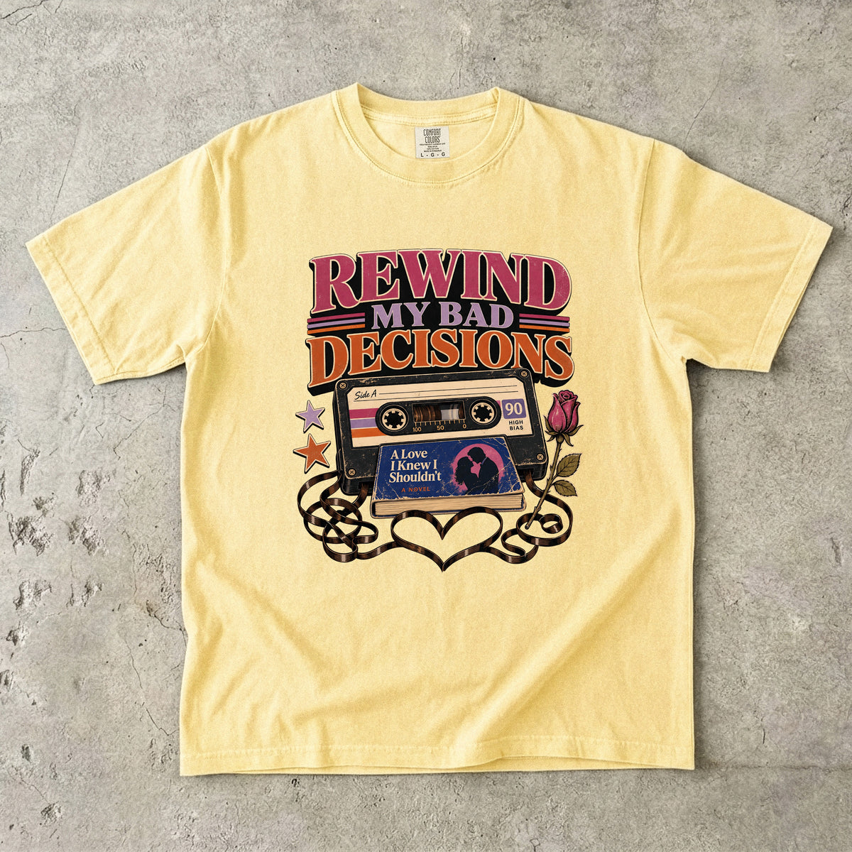 A pale yellow short-sleeve t-shirt laid flat on a textured gray concrete surface. The shirt features a retro graphic in the center with the text 'REWIND MY BAD DECISIONS' above a vintage cassette tape, a pink rose, and black swirl patterns.