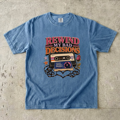 A light blue short-sleeve graphic t-shirt laid flat on a textured concrete surface. The graphic features retro 90s cassette tapes, stars, and the text 'REWIND MY BAD DECISIONS' in pink, orange, and yellow.