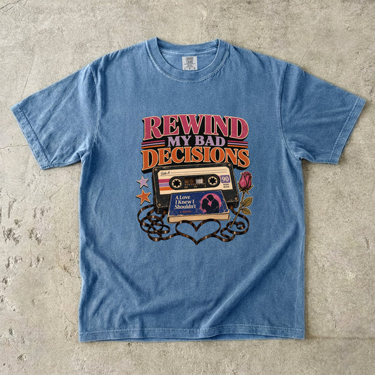 A light blue short-sleeve graphic t-shirt laid flat on a textured concrete surface. The graphic features retro 90s cassette tapes, stars, and the text 'REWIND MY BAD DECISIONS' in pink, orange, and yellow.