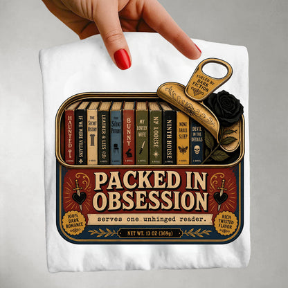 A white T-shirt folded and held by a hand with red nail polish, featuring a large vintage-style book-themed graphic that reads 'Packed in Obsession' with illustrated books, a gold key, a black rose, and decorative text on a light gray background.