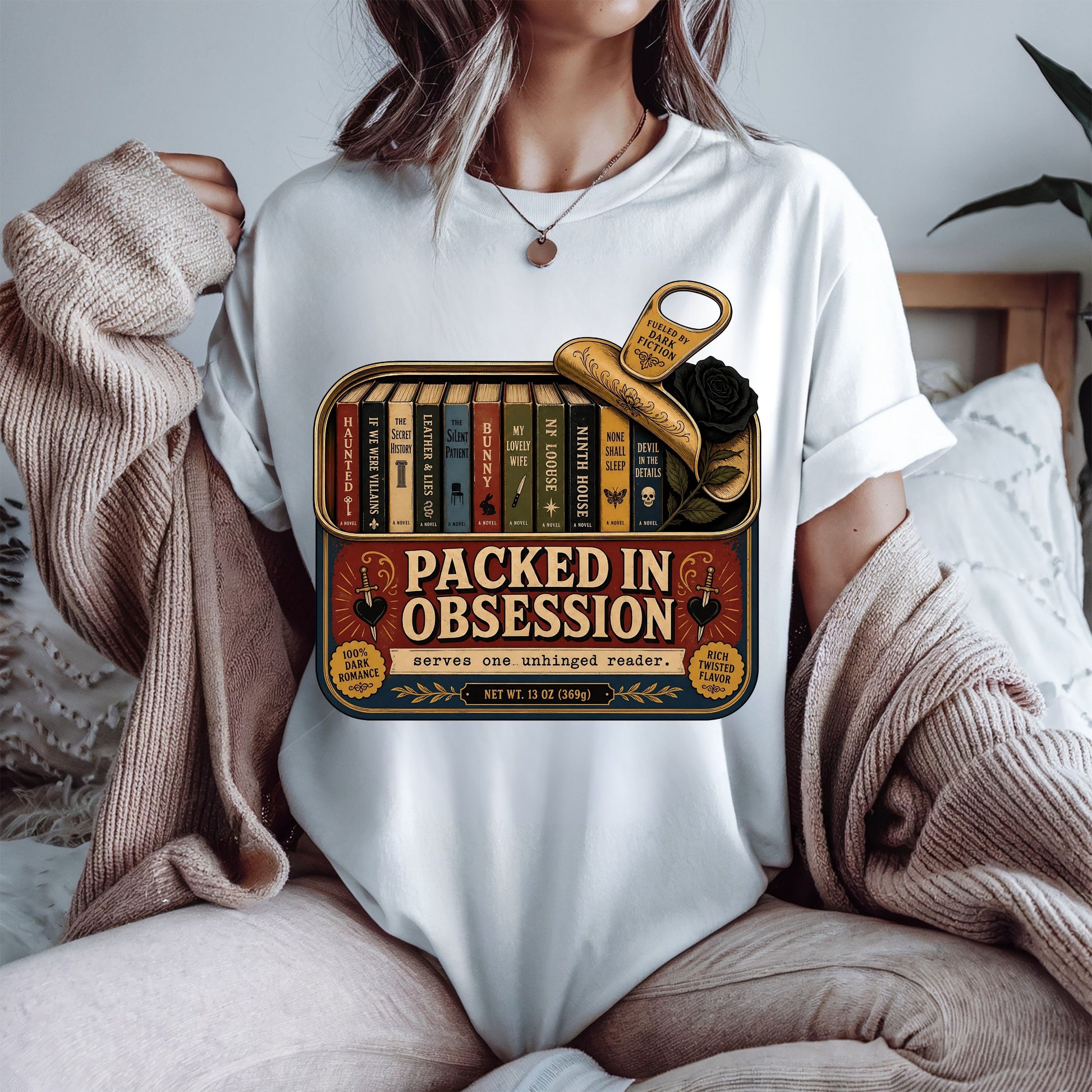 A person wearing a white graphic T-shirt sits on a bed with a beige knit sweater draped off the shoulders. The shirt features a vintage-style book tin graphic with the text 'PACKED IN OBSESSION' and illustrated books, a gold key, and a black rose.